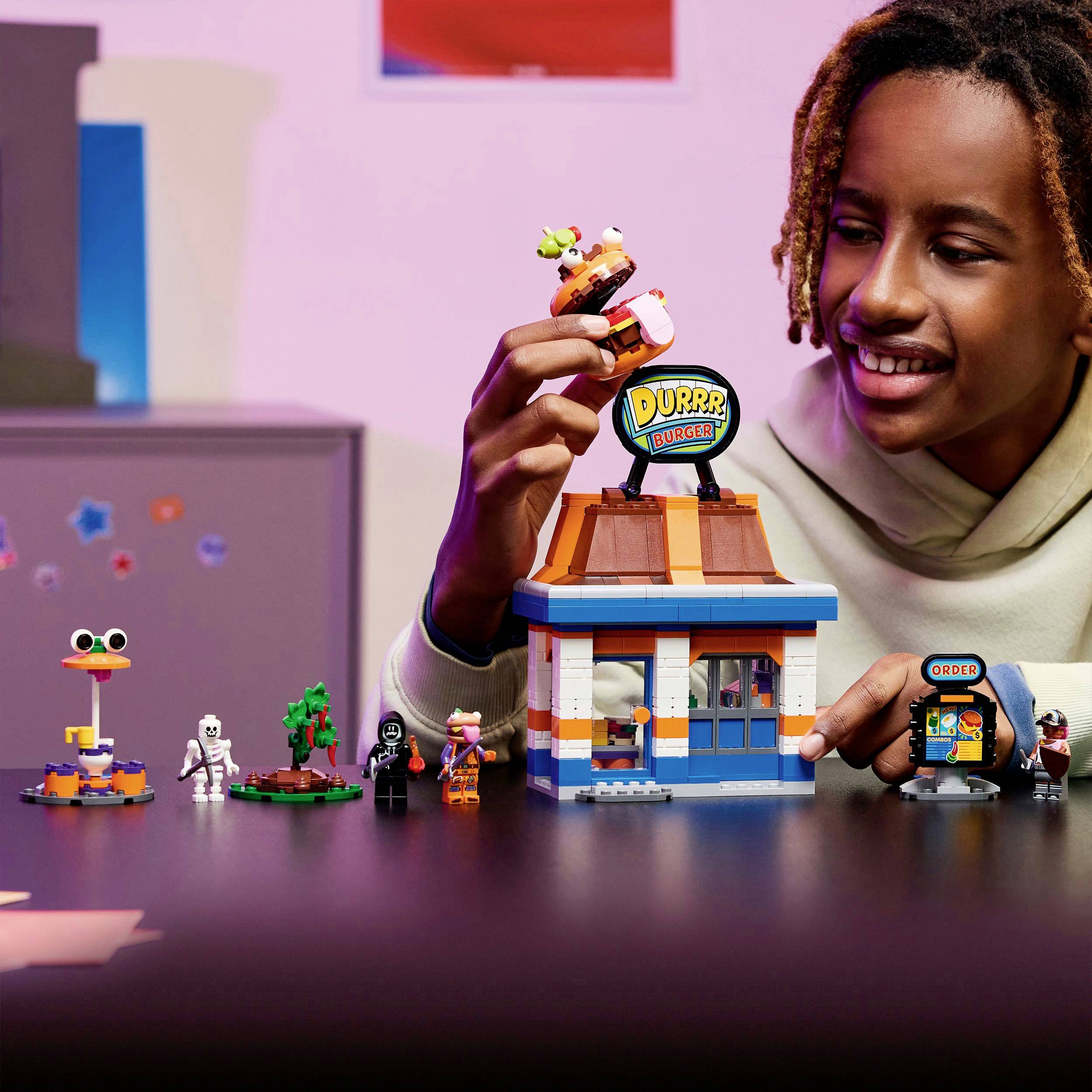 Un bambino gioca con set LEGO, tra cui un modello "Durr Burger" e varie piccole figure. La scena è vivace e colorata.