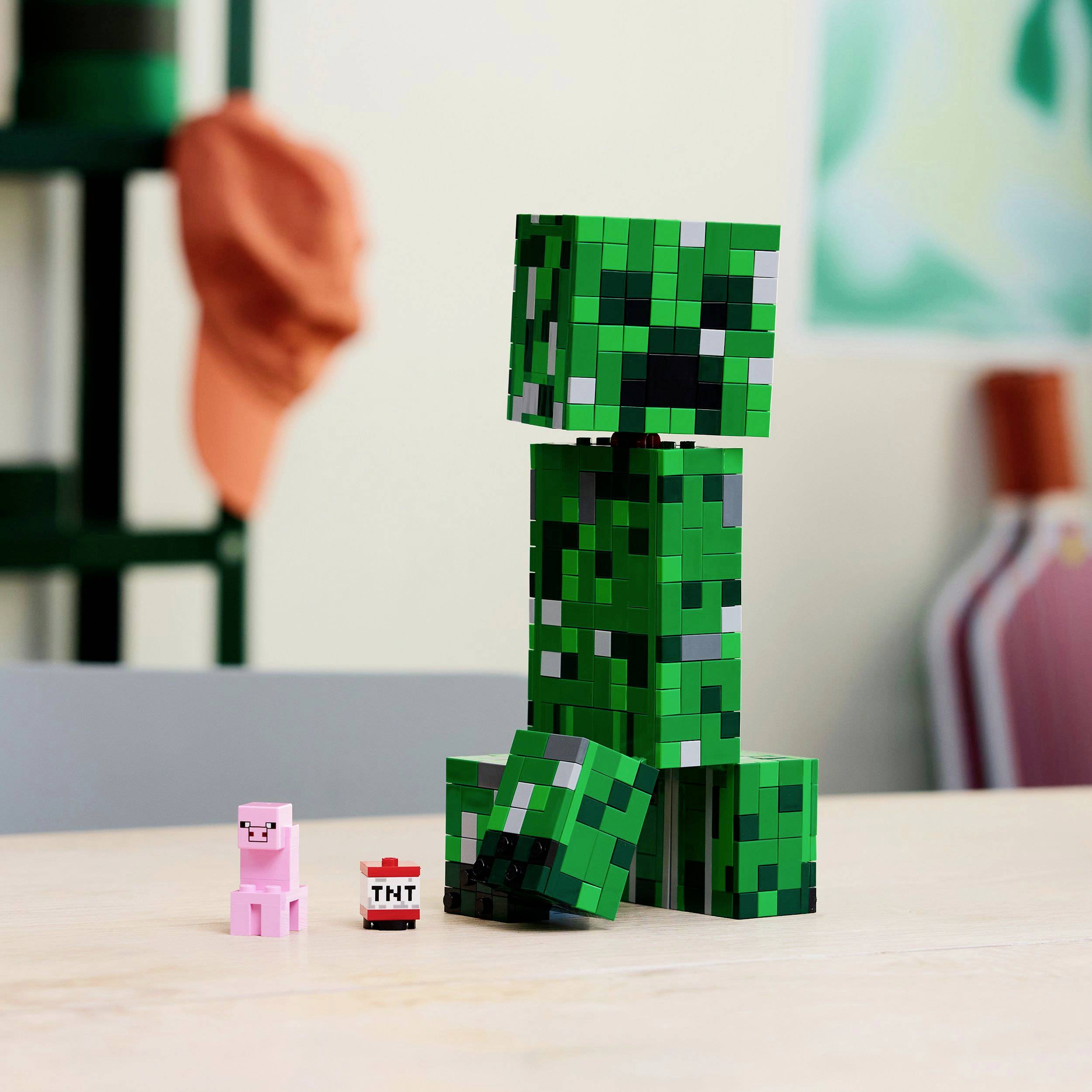 Un Creeper fatto di mattoncini è in piedi su un tavolo, accanto a un maialino e a un blocco di TNT, davanti a uno sfondo sfocato.