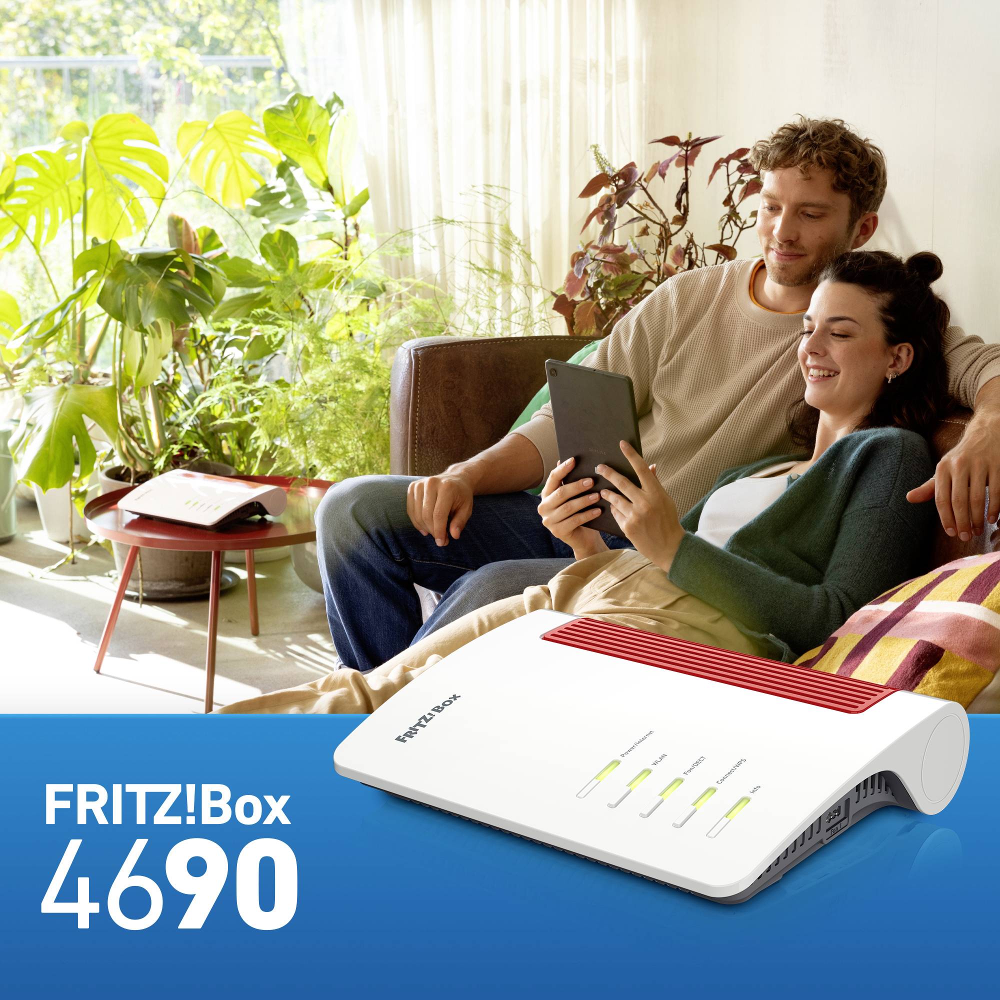 Una coppia sorridente è seduta su un divano e utilizza un tablet. In primo piano è raffigurato un router FRITZ!Box 4690.