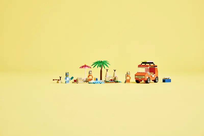 Figure di giocattoli in una scena di spiaggia con personaggi colorati, palma, ombrellone, tavole da surf e un furgone arancione, sullo sfondo giallo.