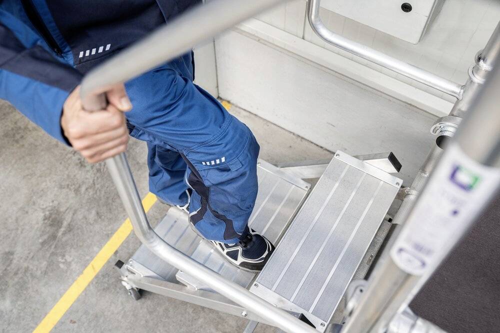 Una persona con indumenti da lavoro blu sta salendo una scala mobile in alluminio. La scala ha gradini antiscivolo.