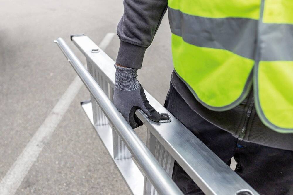Una persona con abiti da lavoro e gilet di sicurezza sta trasportando una scala metallica su una strada. Indossa guanti protettivi.