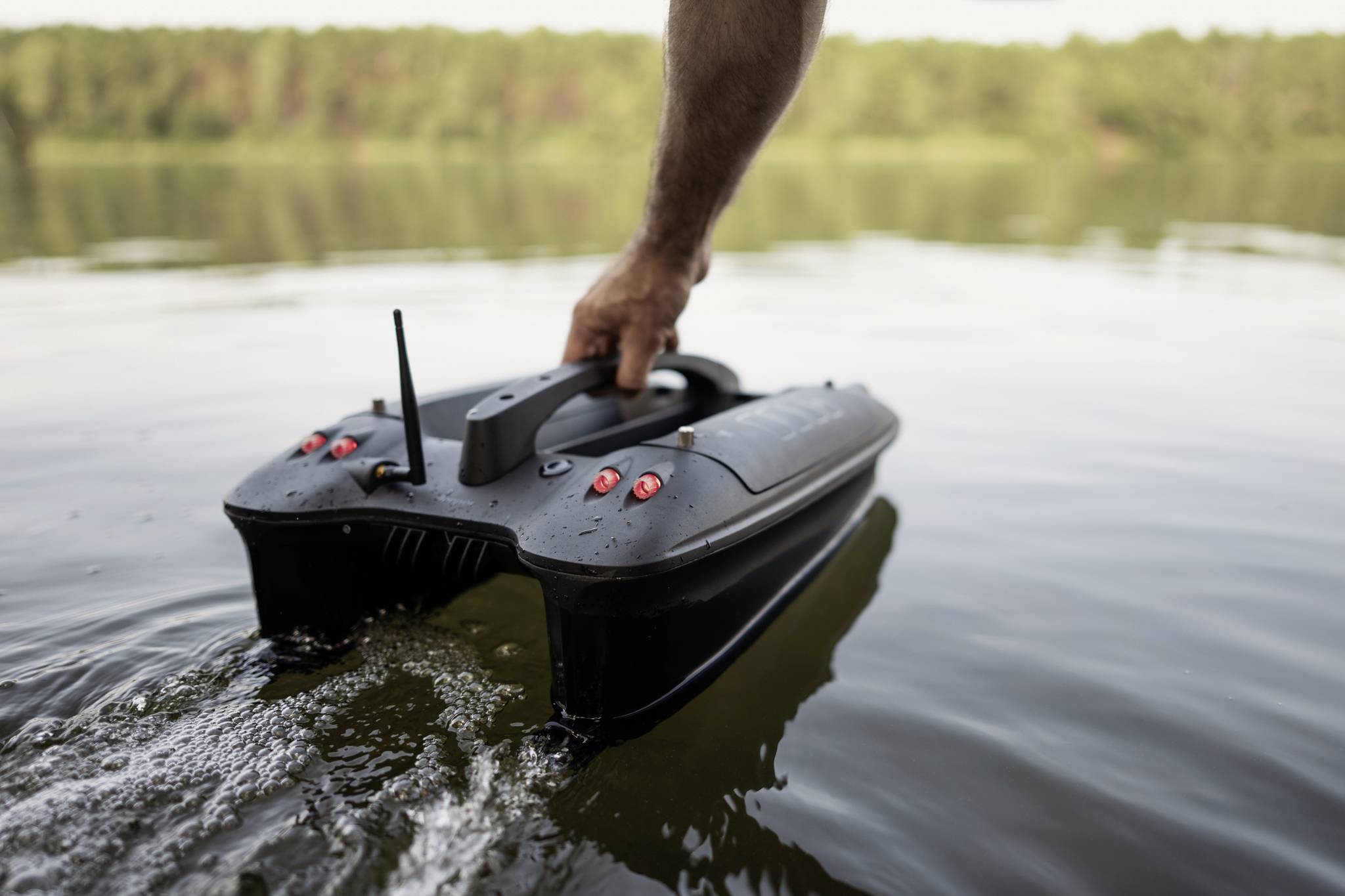 Una barca telecomadata viene guidata da una mano attraverso acque marine calme. Sullo sfondo si intravede una riva boscosa.