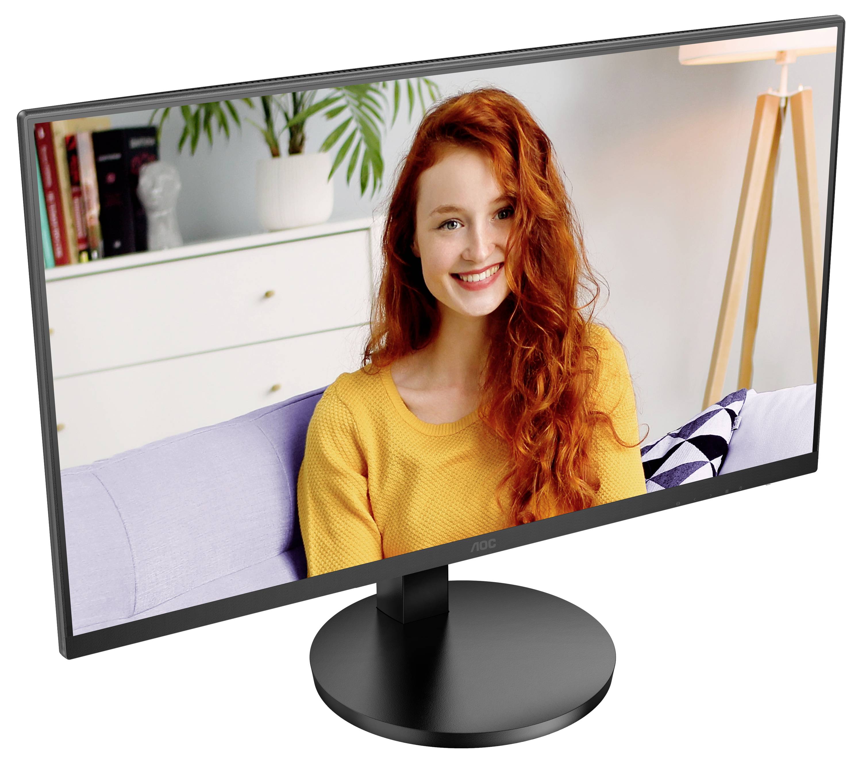 Un monitor di computer mostra una giovane donna con i capelli rossi seduta su un divano, sorridente. Sullo sfondo si vedono piante e libri.