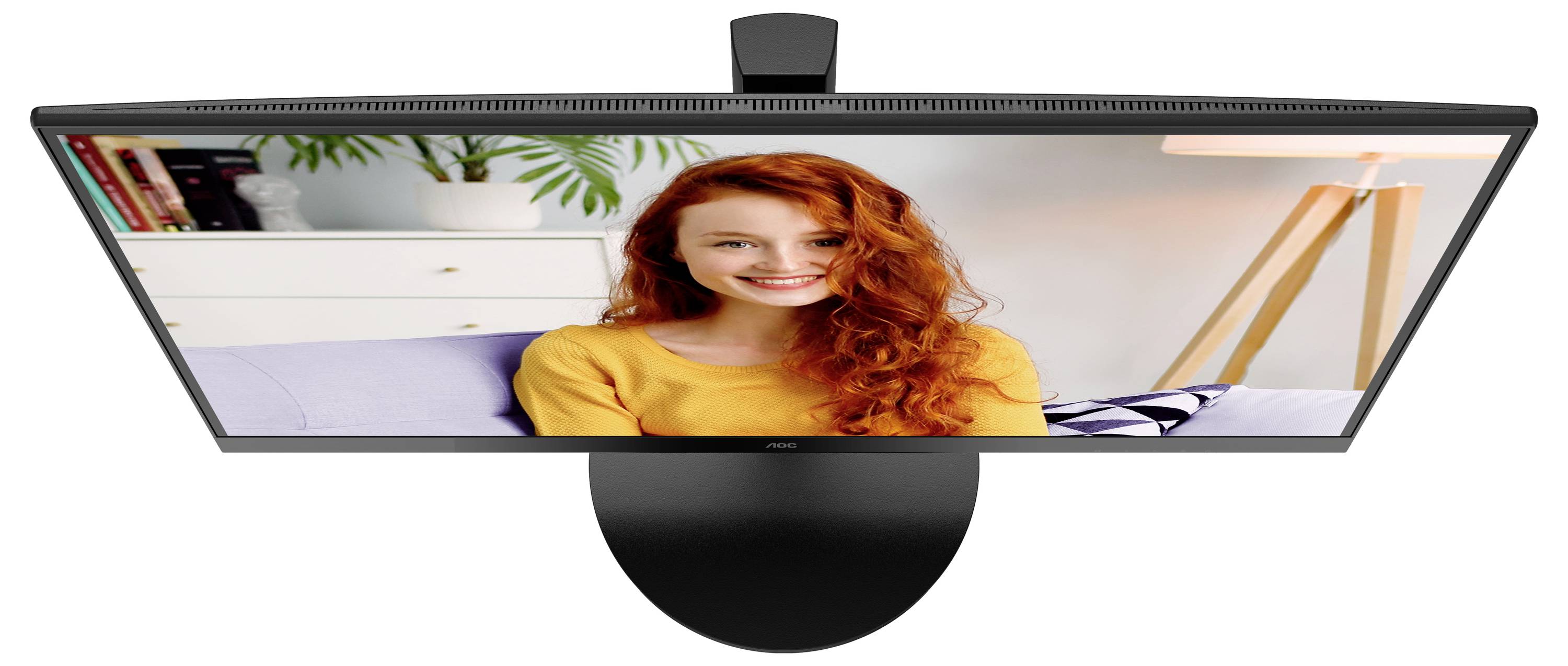 Una persona sorridente con i capelli rossi è seduta su un divano e guarda verso la telecamera. Sullo sfondo ci sono piante, libri e una lampada da terra.
