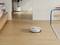 Un robot aspirapolvere bianco su un pavimento di legno chiaro in salotto, accanto a una sedia blu e a un vaso per piante.