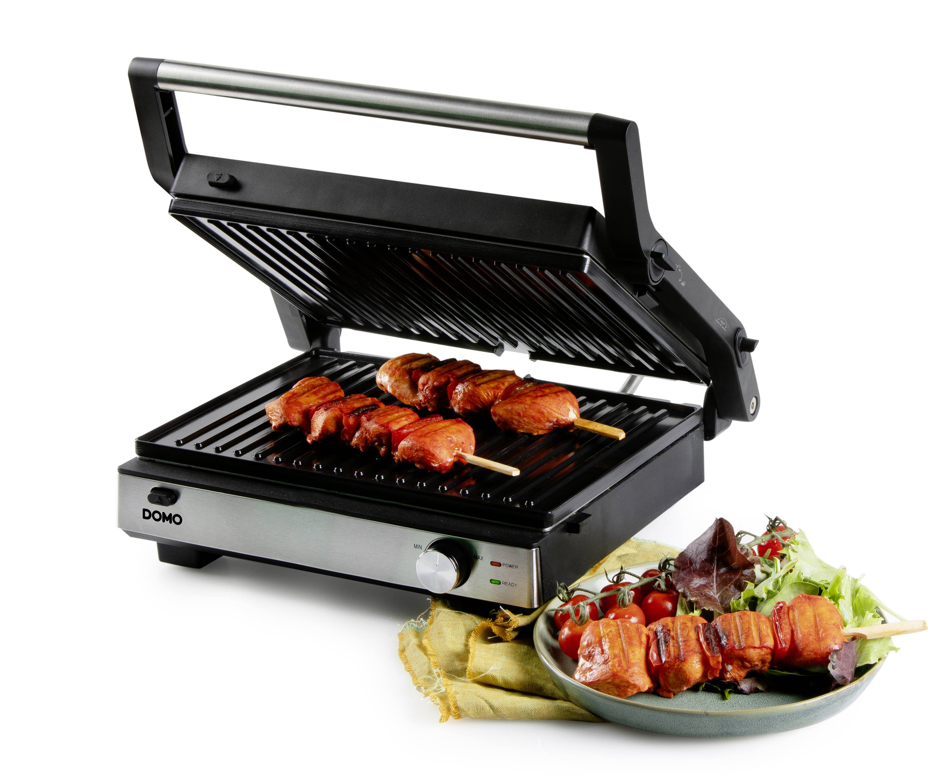 Un barbecue elettrico con il coperchio aperto sta grigliando due spiedini di carne. Accanto c'è un piatto con foglie di insalata fresche e pomodori.