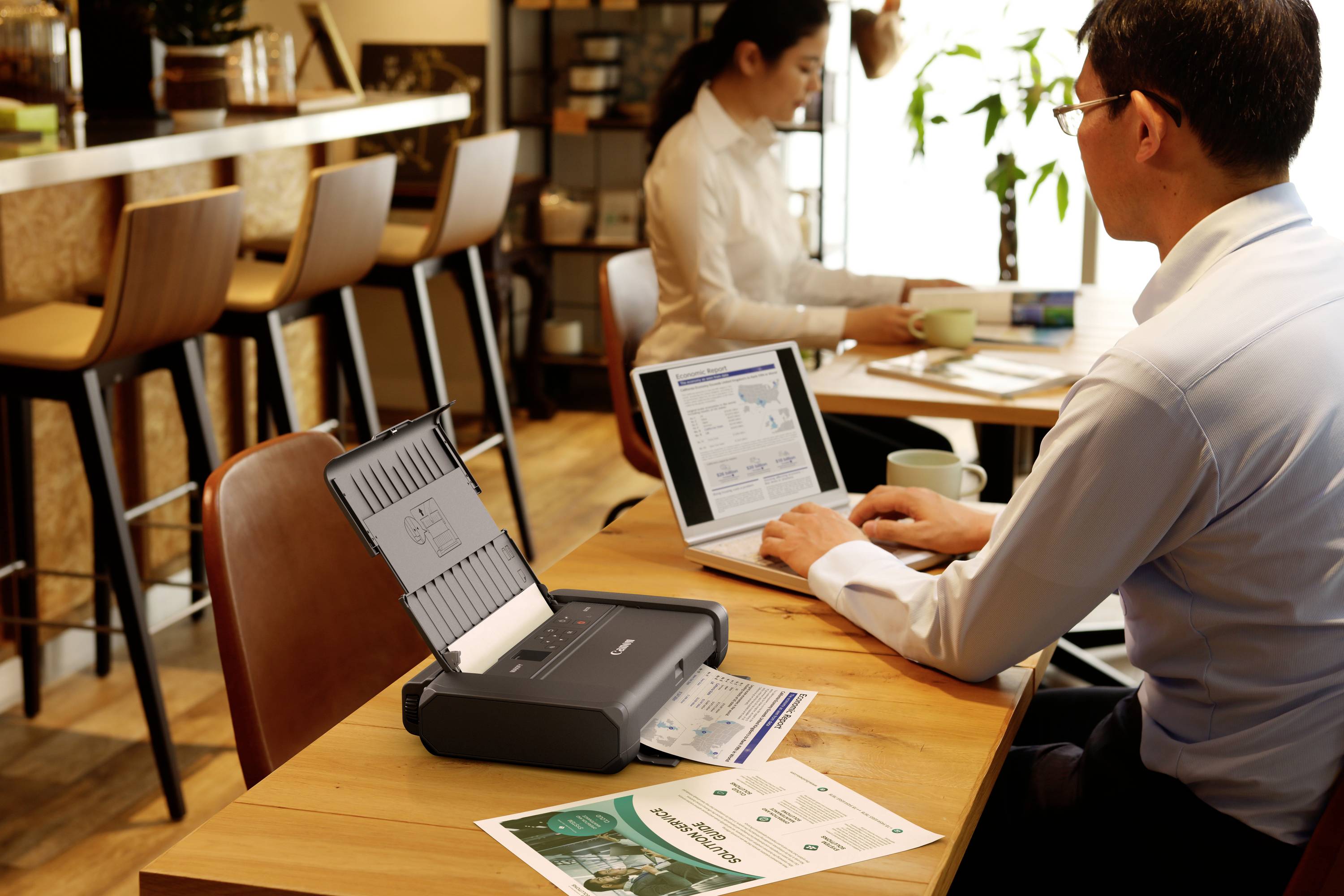 Un uomo lavora su un laptop in un caffè. Davanti a lui c'è una stampante compatta che sta stampando documenti. Sullo sfondo è seduta una donna.