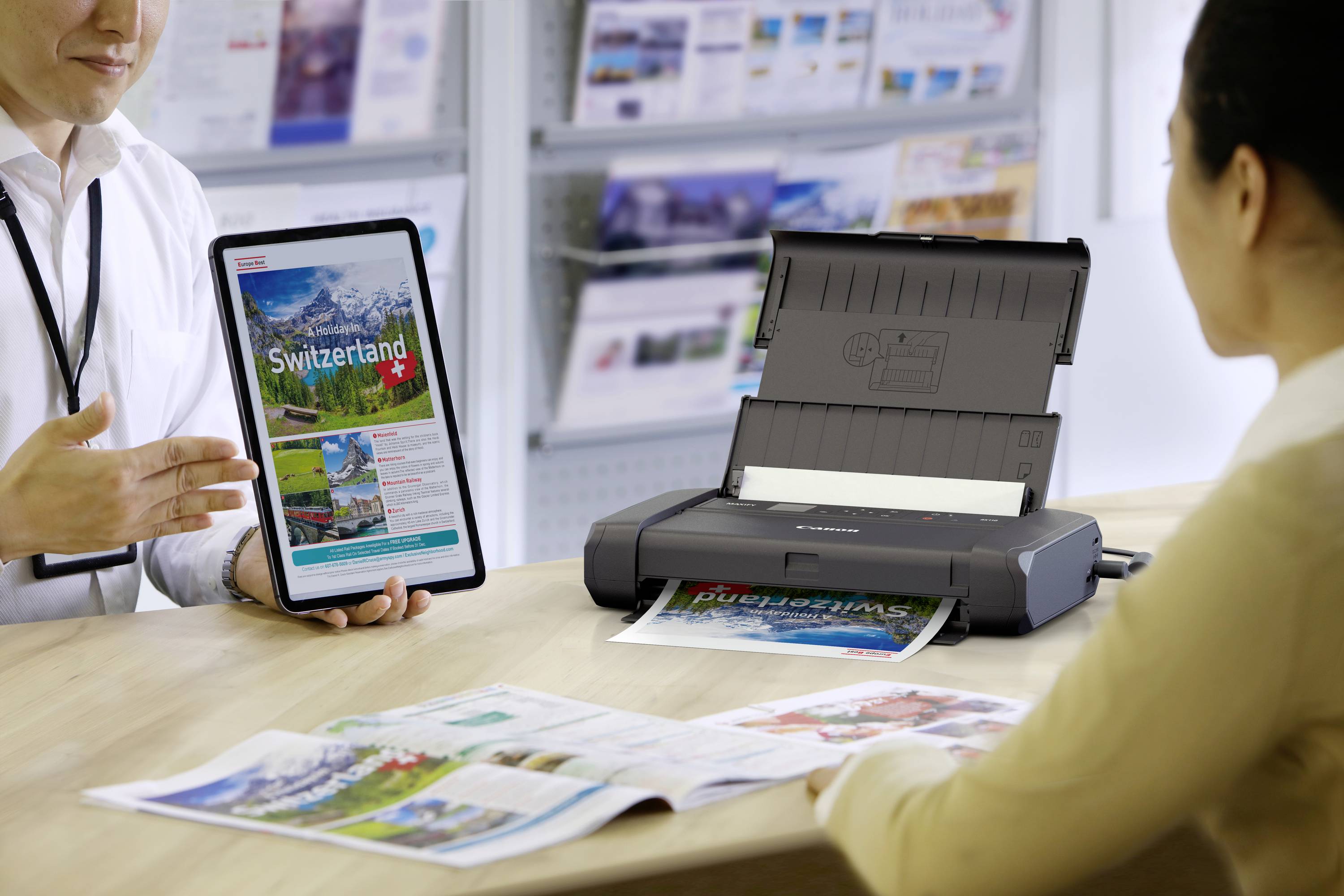 Una persona mostra a un'altra un tablet con la scritta 'Switzerland', mentre una stampante sta stampando un documento a colori.