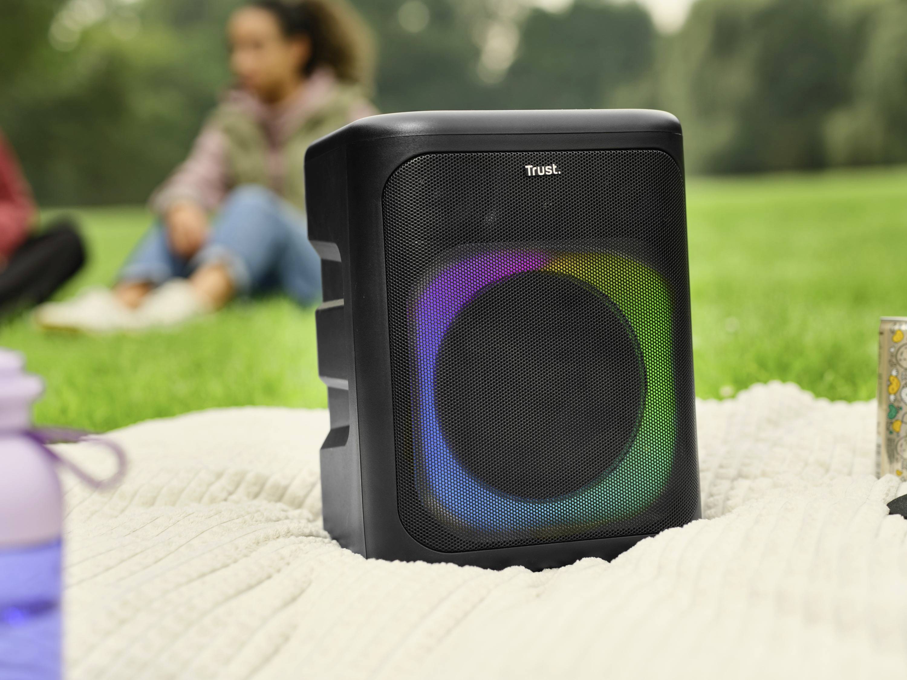 Altoparlante portatile con luci LED luminose poggia su una coperta nel parco. Sullo sfondo, persone sfocate che si rilassano.