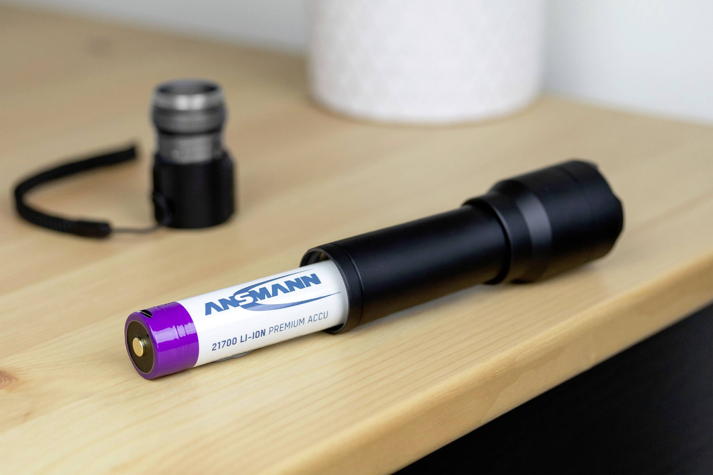 'Ansmann 21700 Li-Ion Battery' lies next to a black flashlight on a wooden table. A blurry additional component is visible in the background.