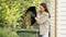 Una donna svuota un contenitore con rifiuti da giardino in un bidone verde, circondataà da piante e con un edificio sullo sfondo.