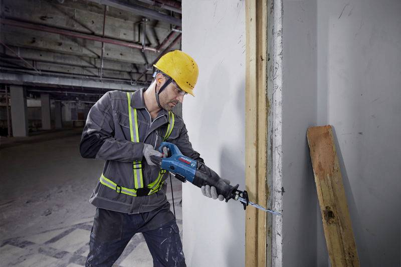 Un operaio edile con casco giallo sta lavorando con una sega elettrica su un pilastro di cemento in un cantiere. Giubbotto di sicurezza visibile.