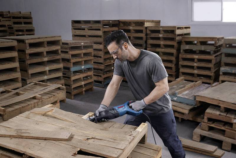 Un uomo con occhiali protettivi e guanti sta segando un pallet con una sega Blaumann in un magazzino pieno di pallet impilati.