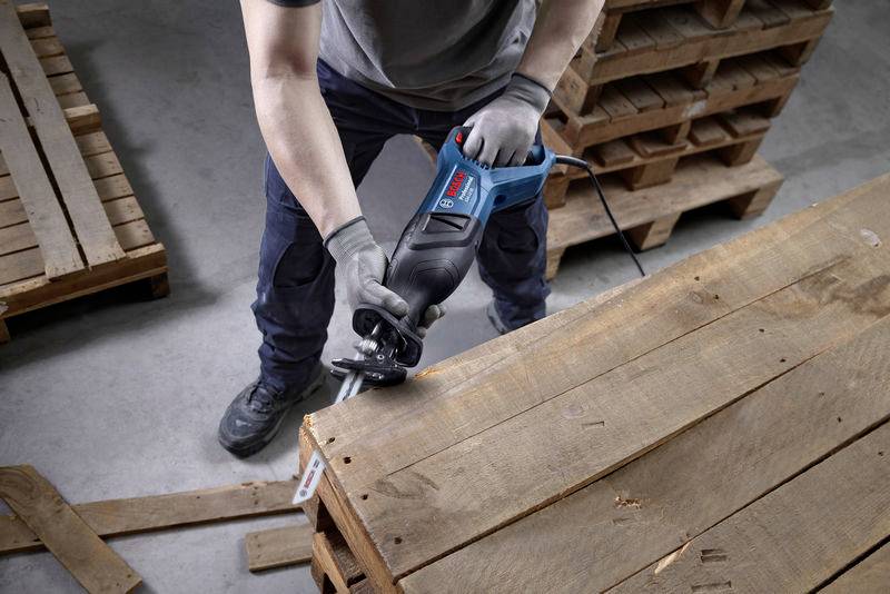 Una persona sta utilizzando una sega elettrica per tagliare un pallet di legno. Sullo sfondo sono visibili altri pallet di legno.