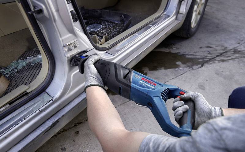 Una persona usa una sega elettrica per rimuovere una portiera dell'auto. Indossa guanti di sicurezza. Pezzi di vetro sono sparsi sul pavimento.