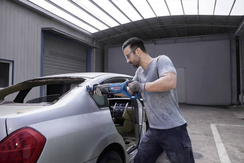 Un uomo con abiti da lavoro sta segando il lunotto posteriore di un'auto argentata con una sega elettrica all'interno di un'officina o di un garage.