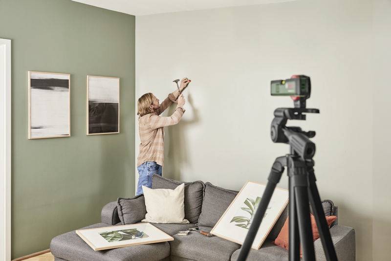 Una persona sta appendendo un quadro al muro in un soggiorno con pareti verde e bianche, un divano grigio e un treppiede con fotocamera.