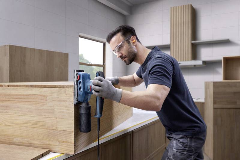 Un uomo sta levigando una superficie di legno con una levigatrice elettrica in un laboratorio. Indossa occhiali protettivi e guanti.