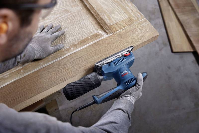 Una persona sta levigando una porta di legno con un levigatrice elettrica blu per smussare i bordi. Sono visibili gli occhiali di protezione e i guanti.