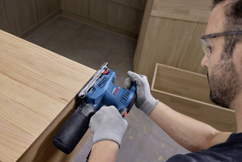 Un uomo sta levigando una superficie di legno con una smerigliatrice blu. Indossa occhiali protettivi e guanti.