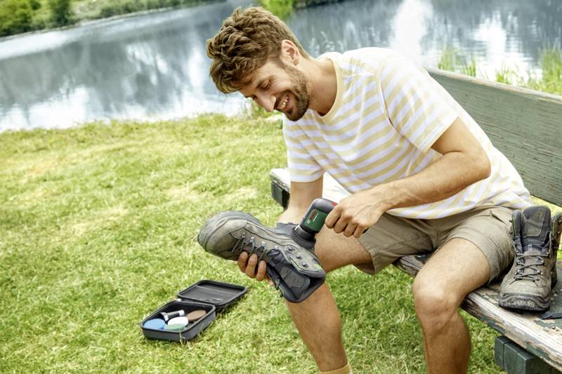 Un uomo è seduto su una panchina vicino al lago e sta pulendo i suoi scarponi da trekking con un dispositivo elettrico. Accanto a lui è posato un kit per la manutenzione delle scarpe aperto.