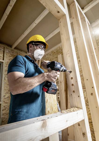 Un operaio edile indossa l'attrezzatura di sicurezza e perfora un telaio in legno con un trapano elettrico in un edificio in costruzione.