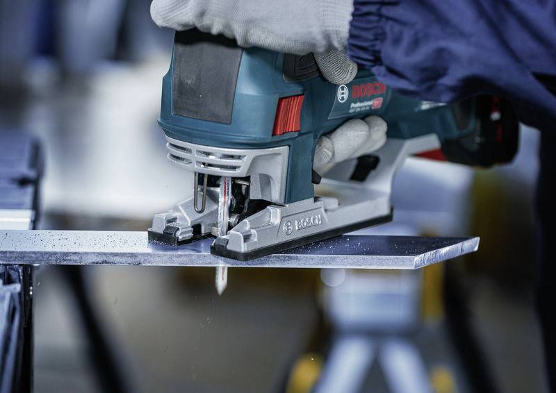 Una persona sta segando un pezzo di metallo con una sega alternativa elettrica. La sega è turchese e la persona indossa guanti da lavoro grigi.