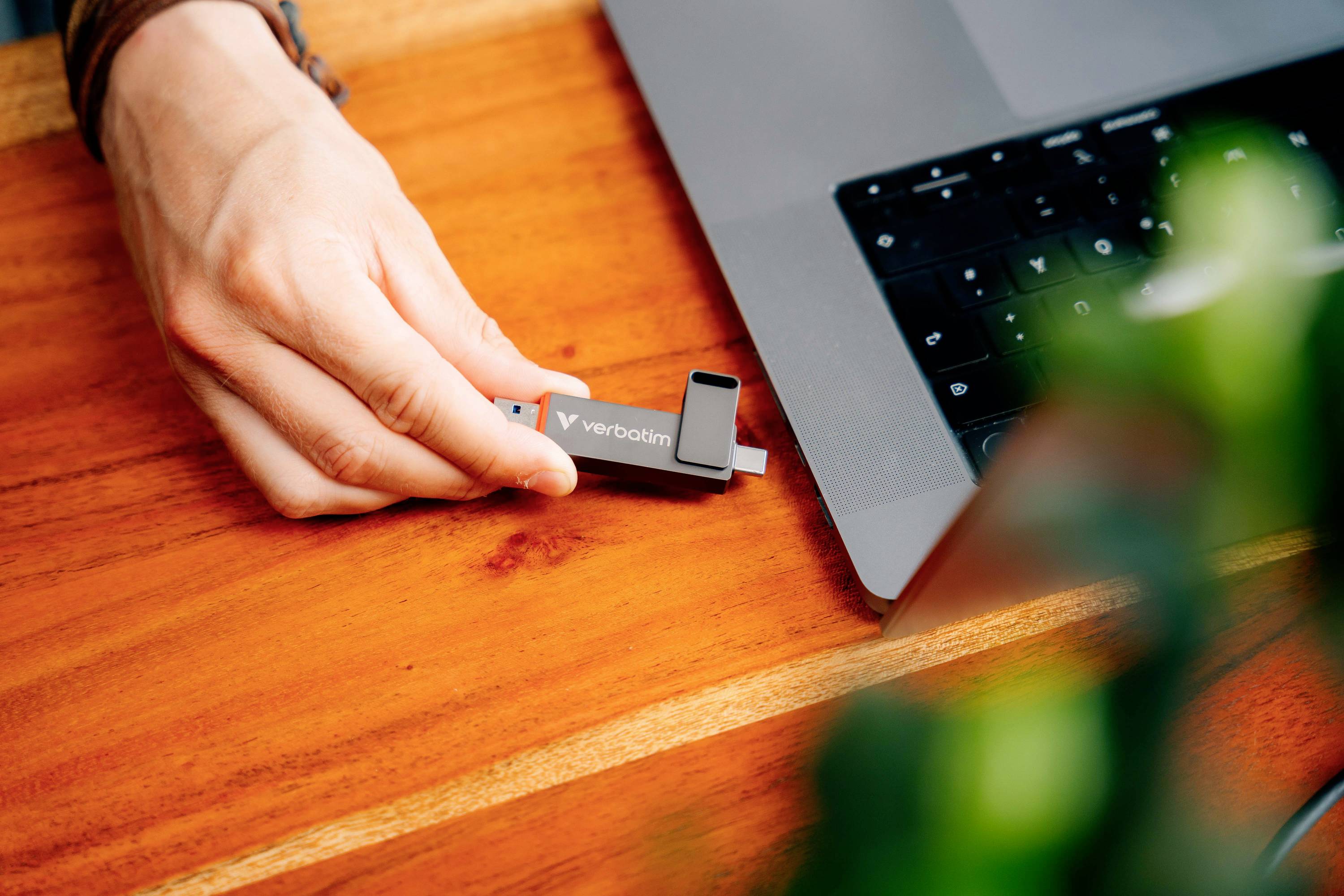 Una mano inserisce una chiavetta USB nera in un laptop su un tavolo di legno. Una foglia verde è sfocata in primo piano.