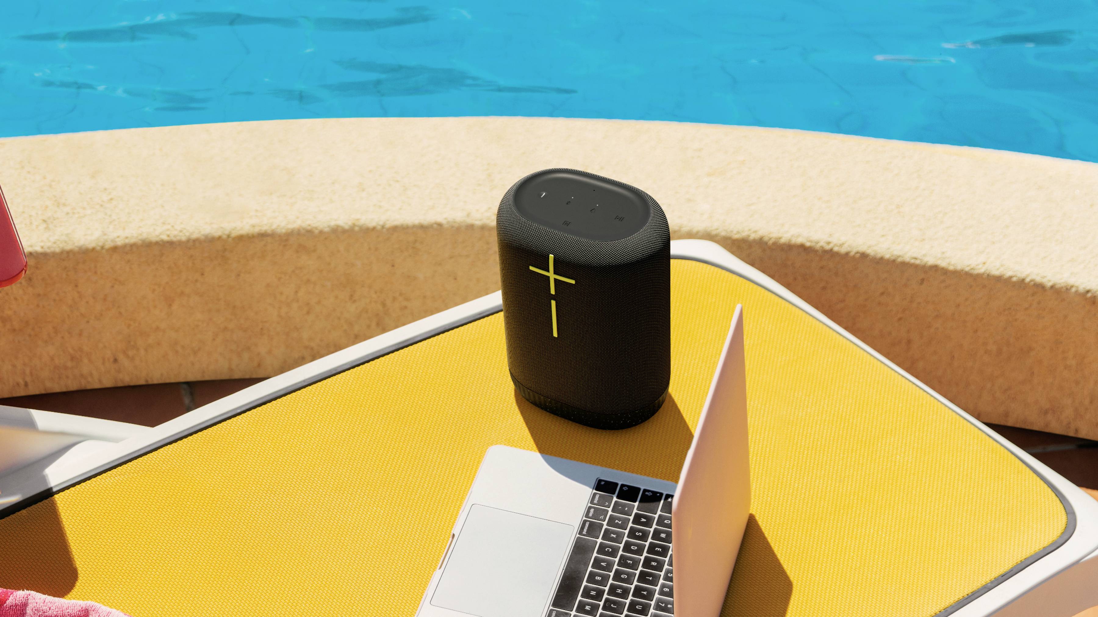 Un laptop e un altoparlante portatile sono posizionati su un lettino giallo sul bordo di una piscina. Il sole illumina la scena.
