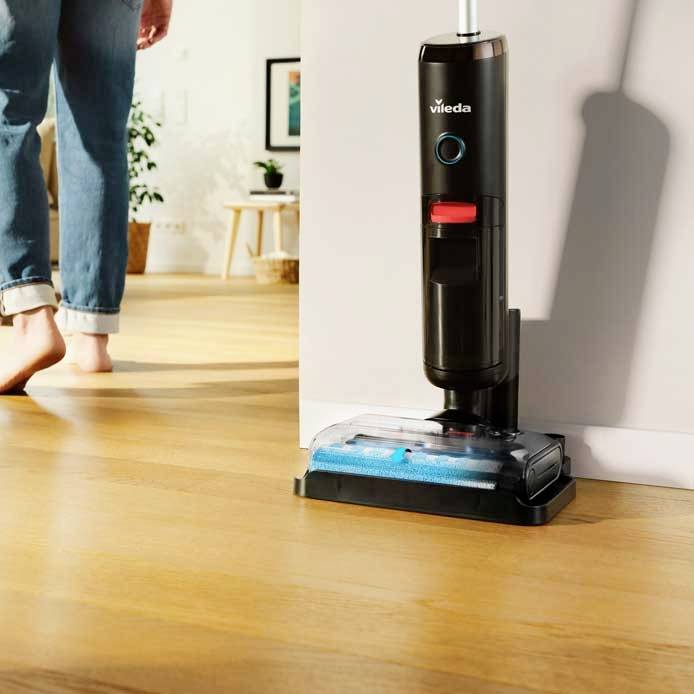 Una persona cammina a piedi nudi su un pavimento in legno, mentre un pulitore per pavimenti Vileda è visibilmente presente nella stanza.