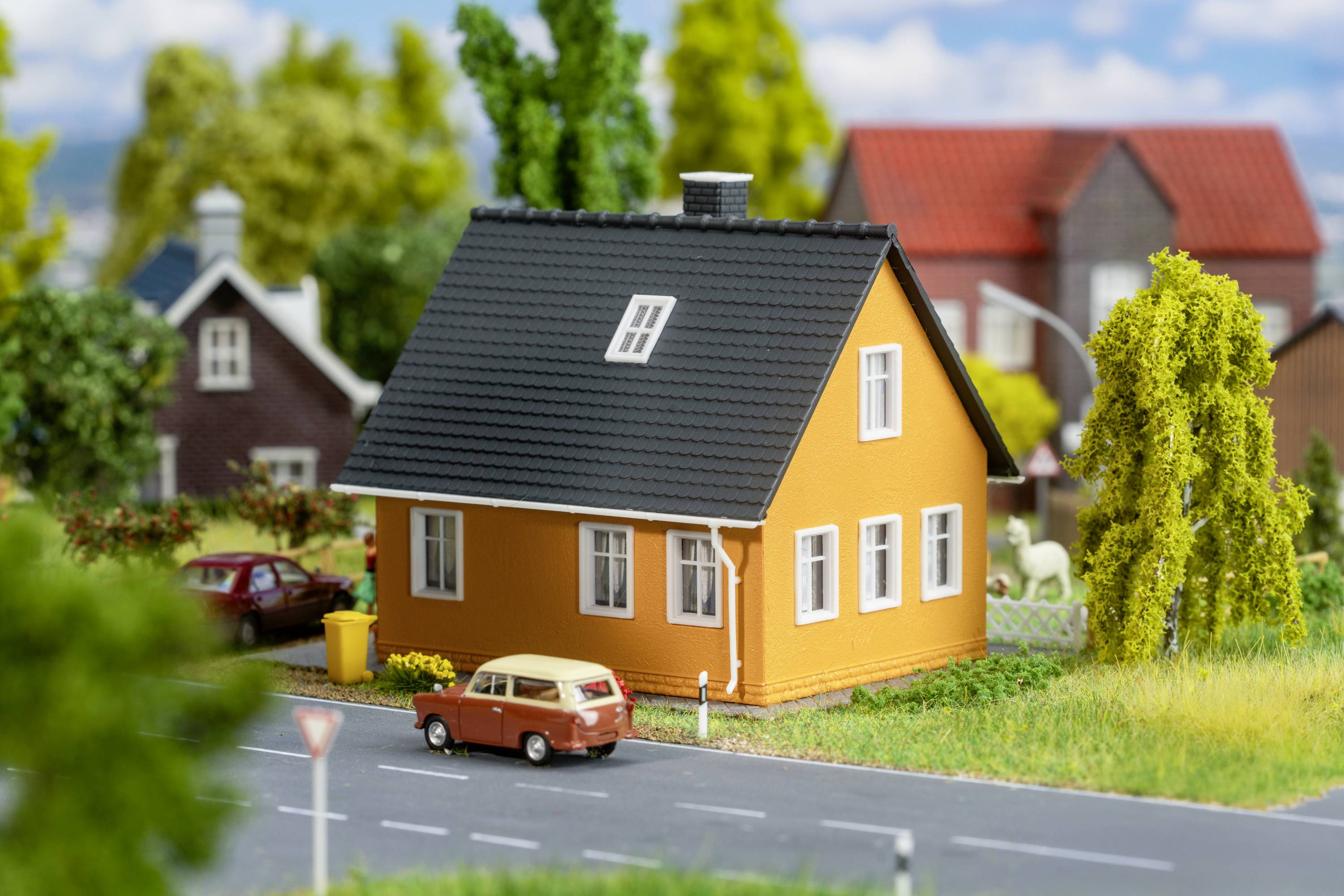 Una casa rossa con un tetto nero e finestre bianche si trova in un paesaggio in miniatura con alberi e una piccola auto.