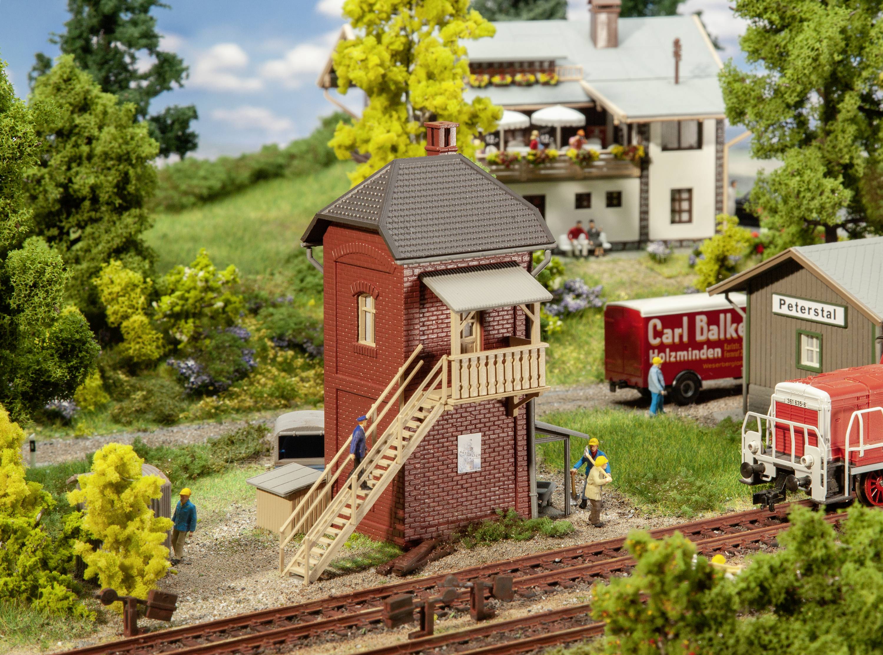 Miniatura di un impianto ferroviario con stazione e locomotiva. Persone animano la scena. Sfondo rurale con alberi e case.