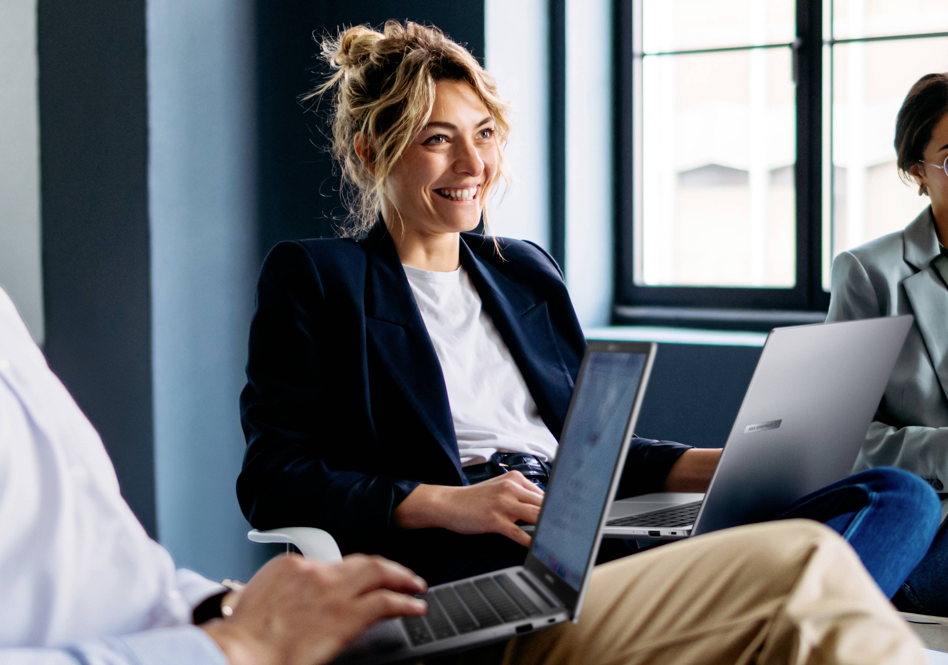 Una persona sorridente siede in un ufficio moderno con laptop, impegnata in un ambiente di lavoro collaborativo.