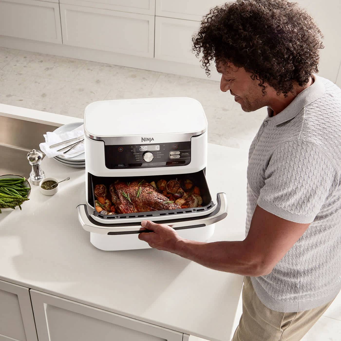 Una persona apre un forno multifunzione bianco Ninja, all'interno del quale è in preparazione un arrosto con verdure, in una cucina moderna.