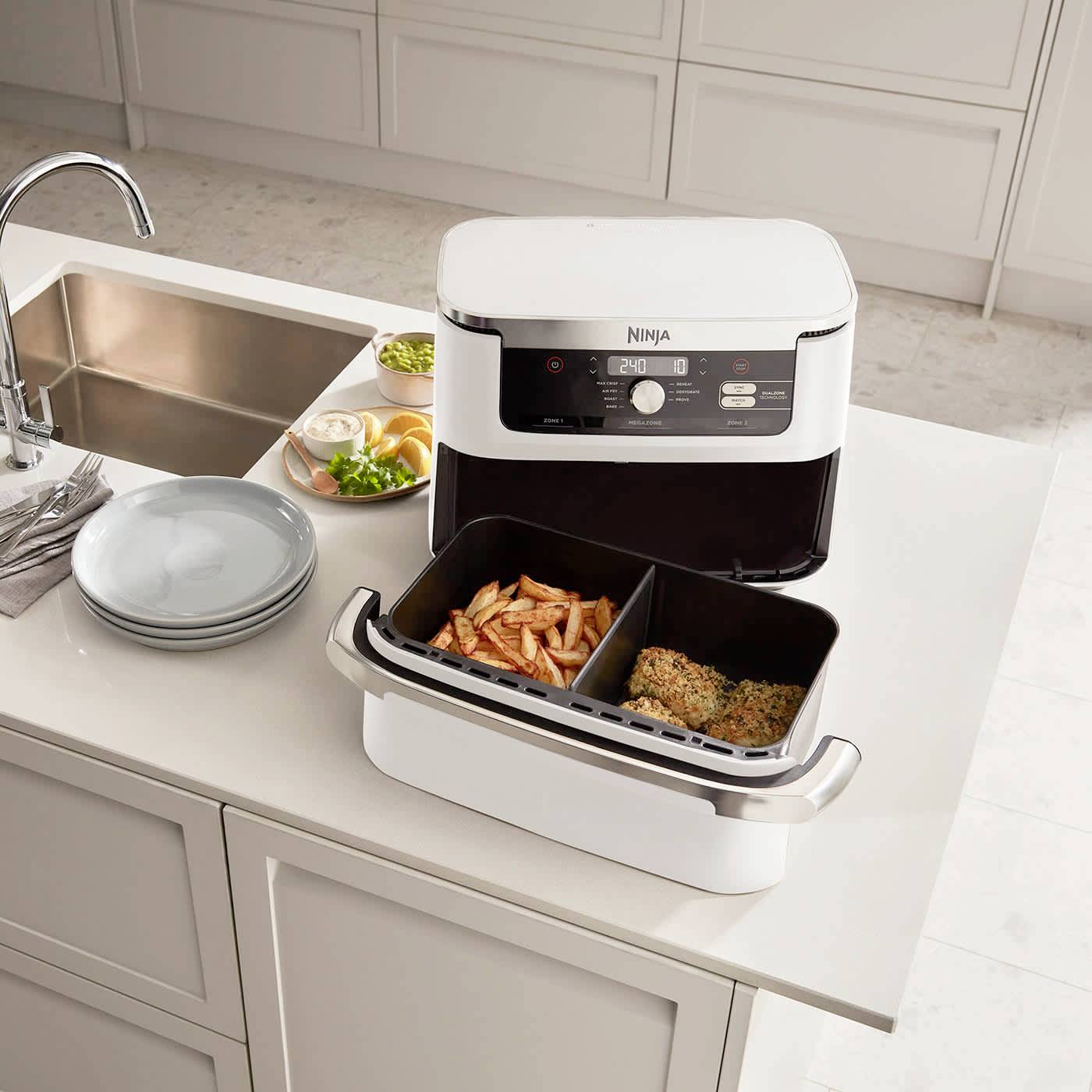 L'immagine mostra una friggitrice ad aria bianca con due vani su un piano di lavoro da cucina. Piatti e verdure sono posizionati accanto ad essa.