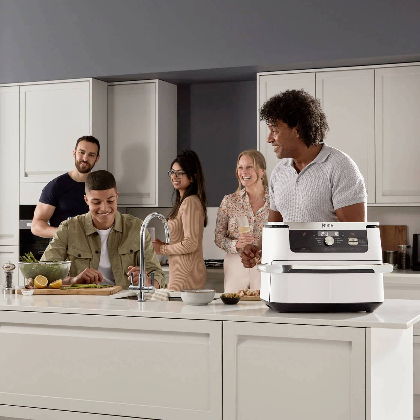 Un gruppo di cinque persone cucina e ride insieme in una cucina moderna. Sul tavolo ci sono utensili da cucina, un colino e degli alimenti.