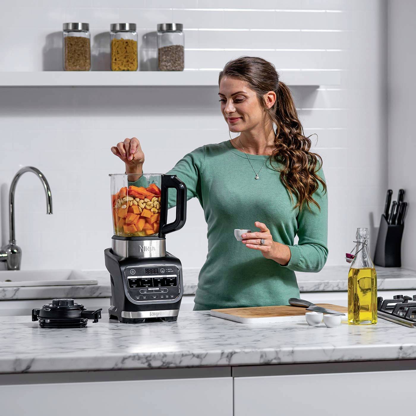 Donna con una blusa verde in piedi in una cucina moderna mentre usa un frullatore con pezzi di verdura. Sullo sfondo ci sono un lavello e utensili da cucina.