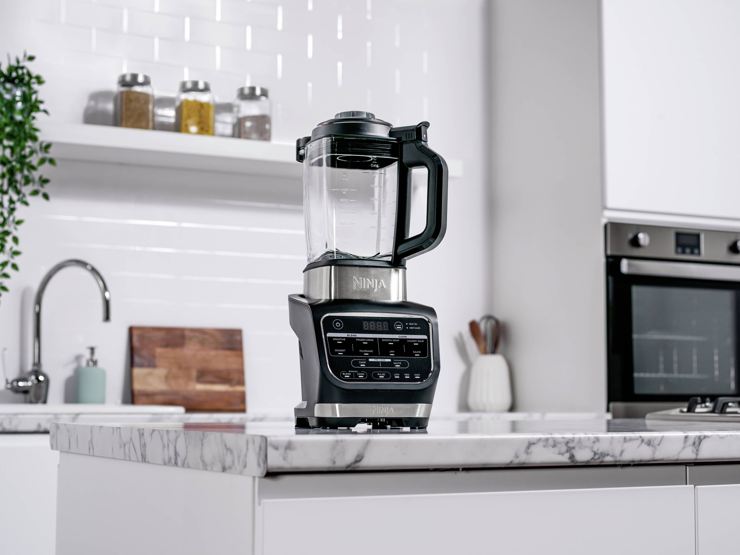 Un mixer argentato è posizionato su un piano di lavoro in marmo. Sullo sfondo si trovano uno scaffale per spezie e moderni elettrodomestici da cucina.