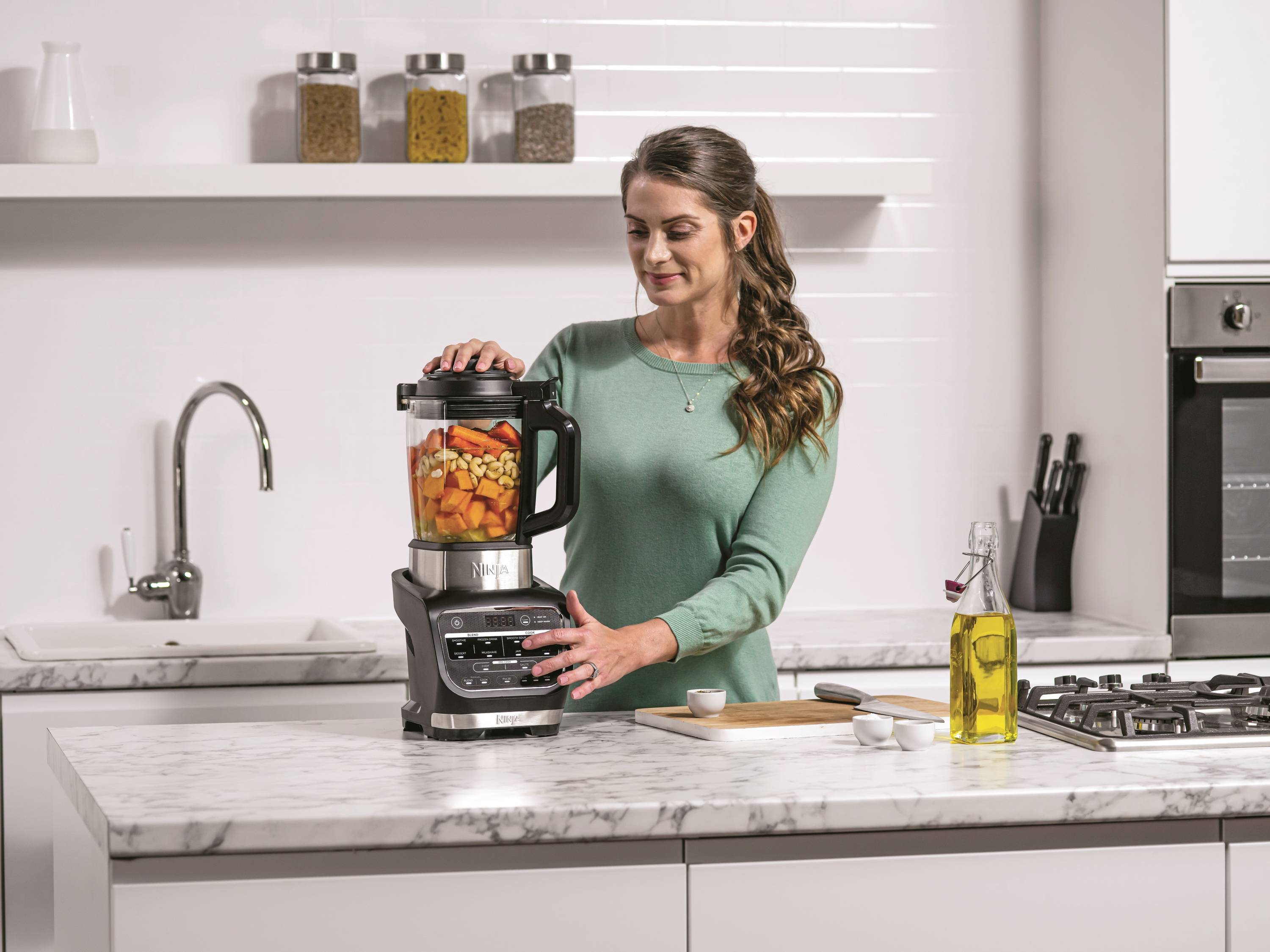 Una donna in cucina sta utilizzando un mixer con gli ingredienti. Accanto a lei ci sono un tagliere, un coltello, barattoli di spezie e una bottiglia di olio.