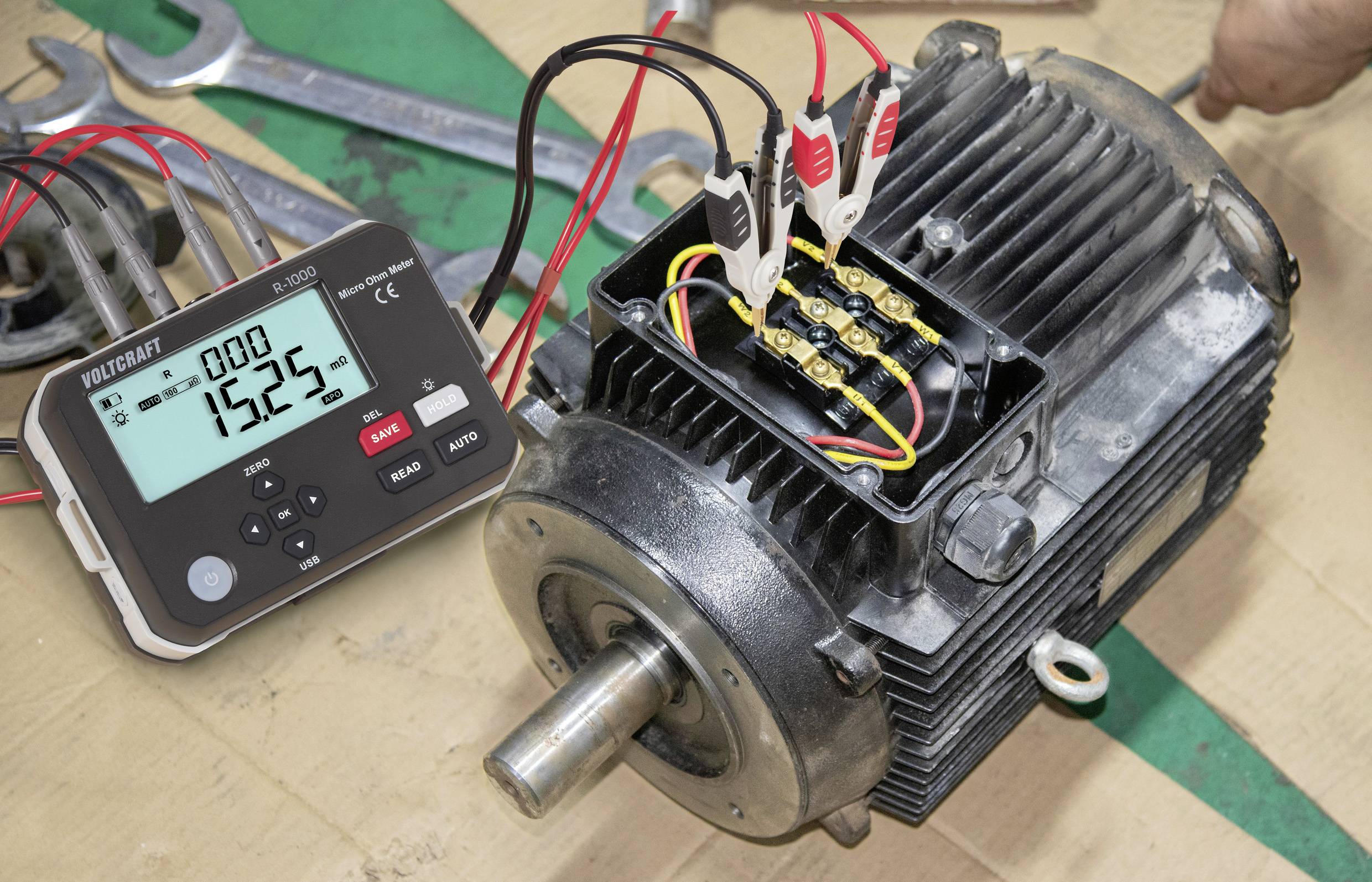 Motore elettrico con fili collegati a un calibratore multifunzione che mostra '1525'. Chiavi inglesi sullo sfondo suggeriscono lavori di manutenzione.