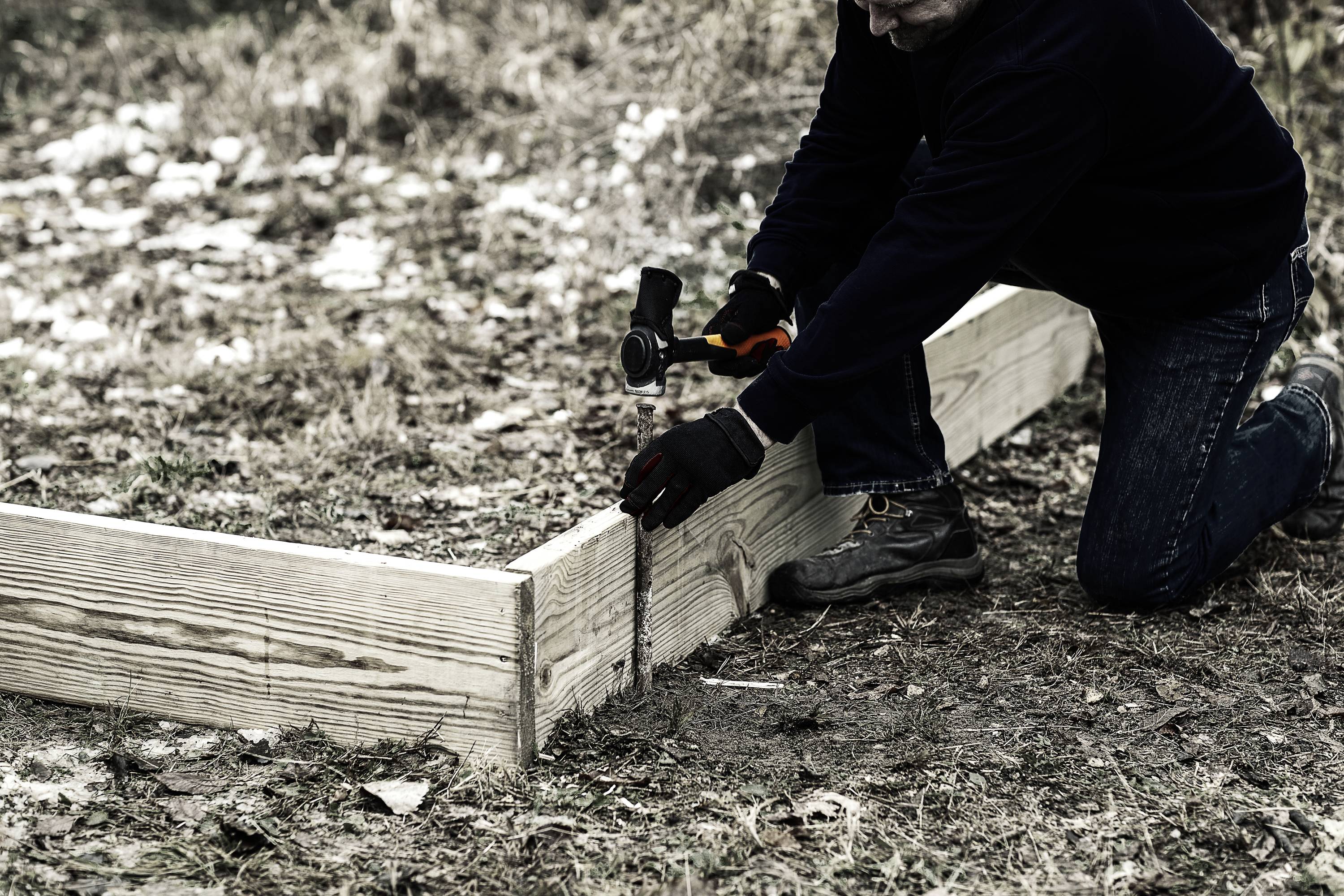 Una persona sta conficcando dei pali nel terreno per fissare tavole di legno. Indossa dei guanti e sta inginocchiata sull'erba.