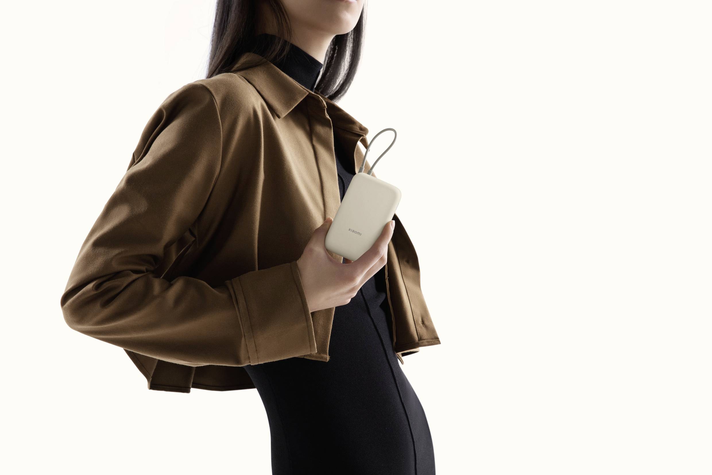 Una persona con una giacca marrone elegante tiene in mano un dispositivo compatto e rettangolare con un'ansa, in piedi davanti a uno sfondo neutro.