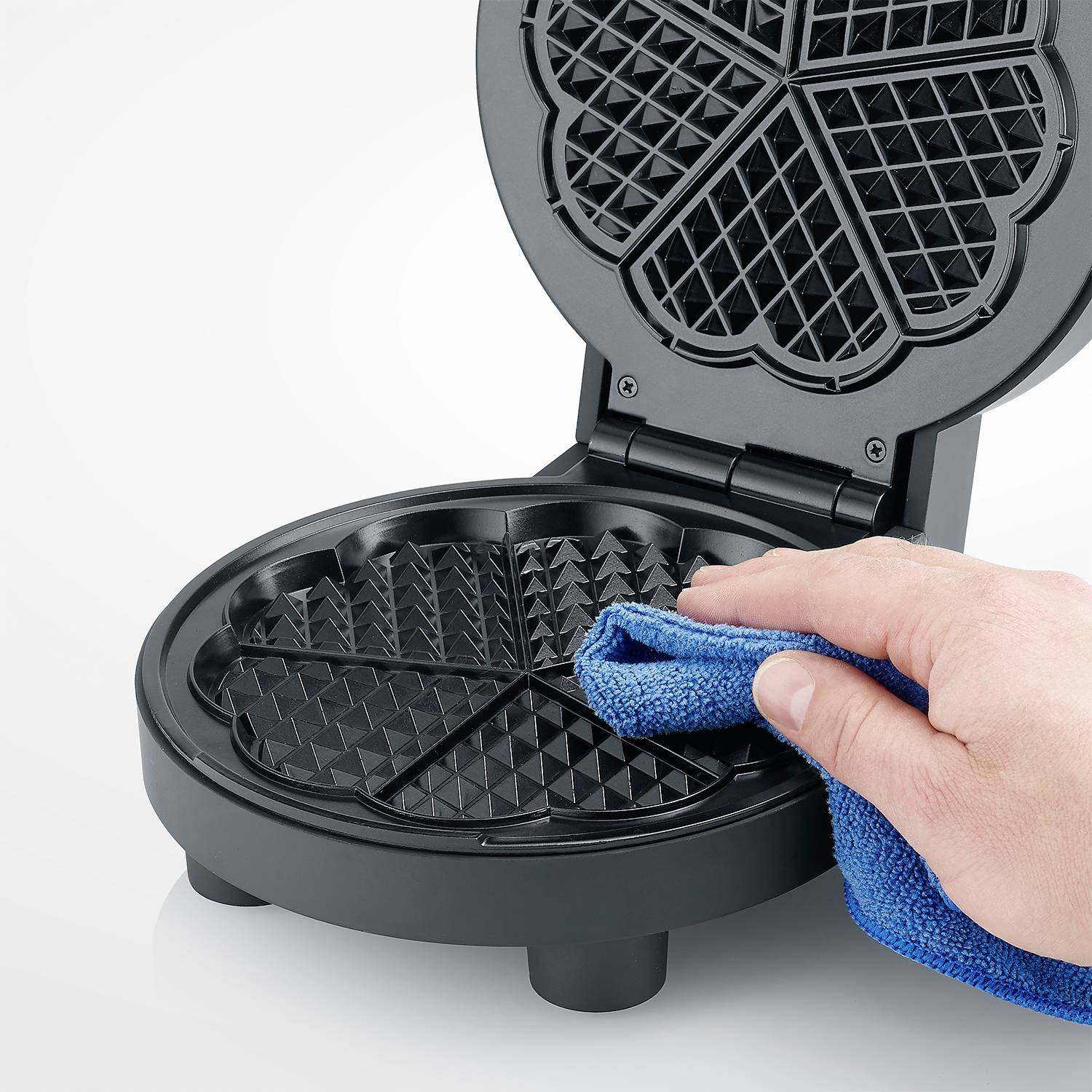 Una mano sta pulendo l'interno di una macchina per waffle con un panno blu. L'apparecchio è aperto e mostra la sua superficie antiaderente nera.