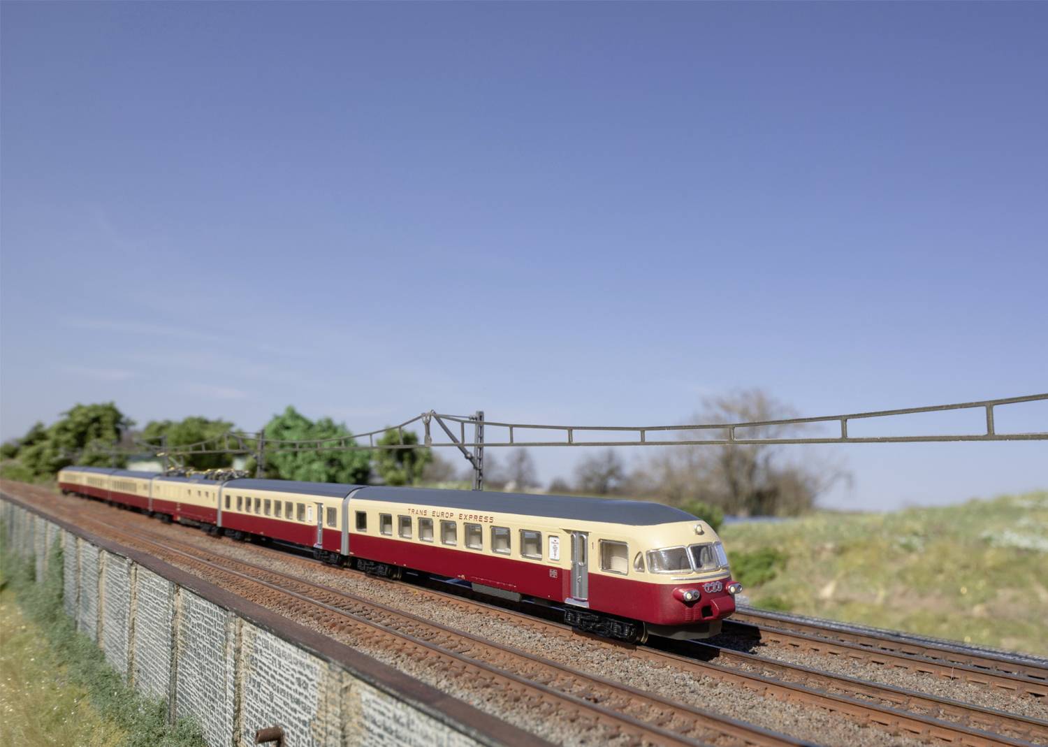 Modello di treno su un percorso ferroviario in miniatura con pali elettrici, circondato da un paesaggio con alberi e un cielo limpido sullo sfondo.