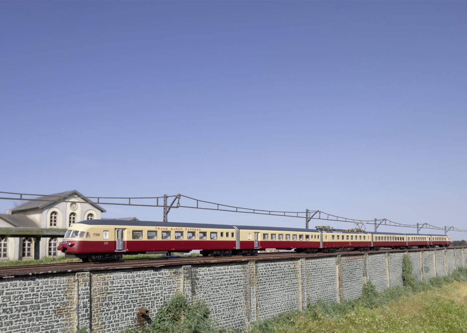 Un treno rosso percorre un binario sopraelevato accanto a un edificio e sotto un cielo blu limpido.