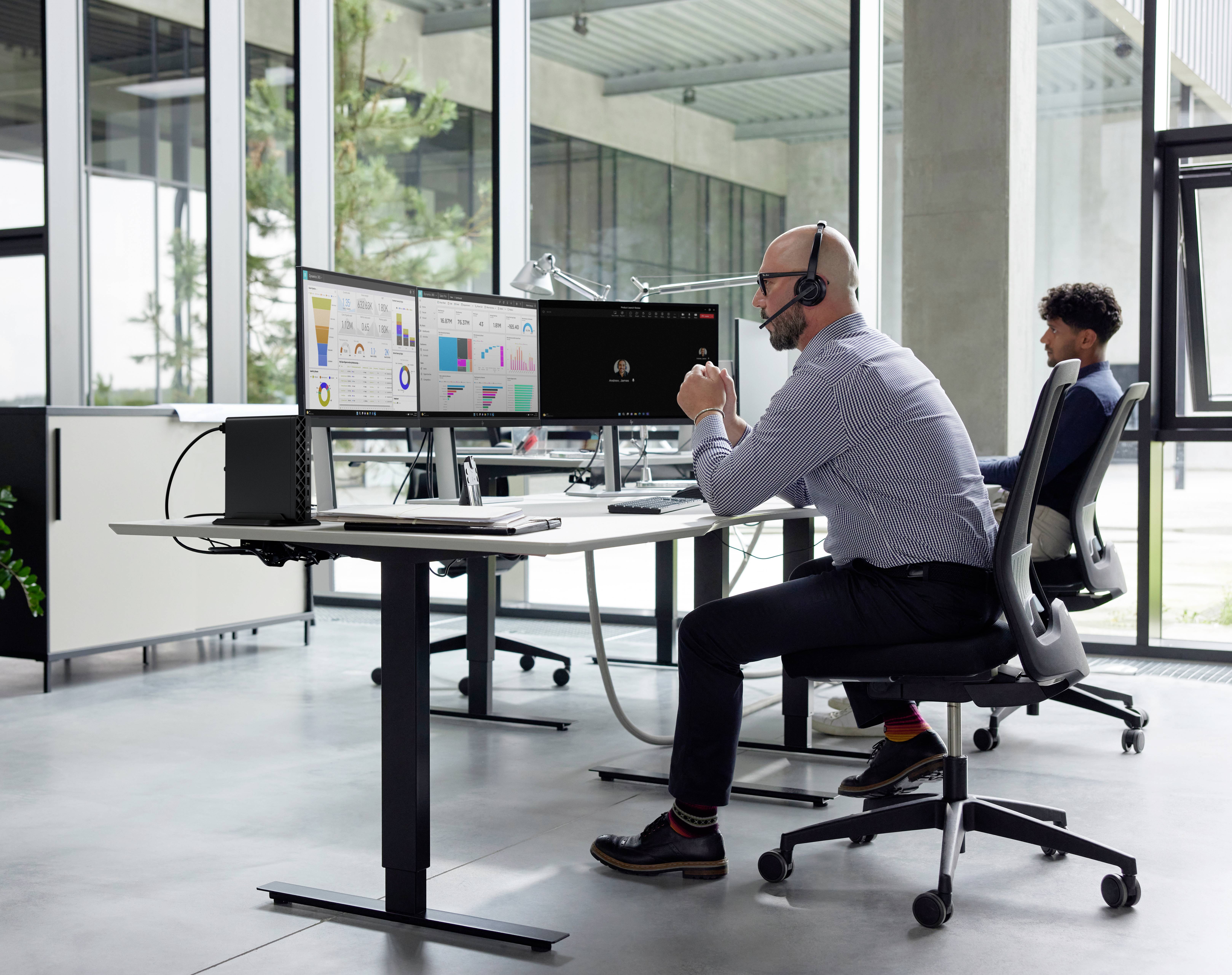 Un uomo con un auricolare lavora a una scrivania con più monitor che mostrano grafici, mentre un'altra persona è seduta a una scrivania vicina in un ufficio.