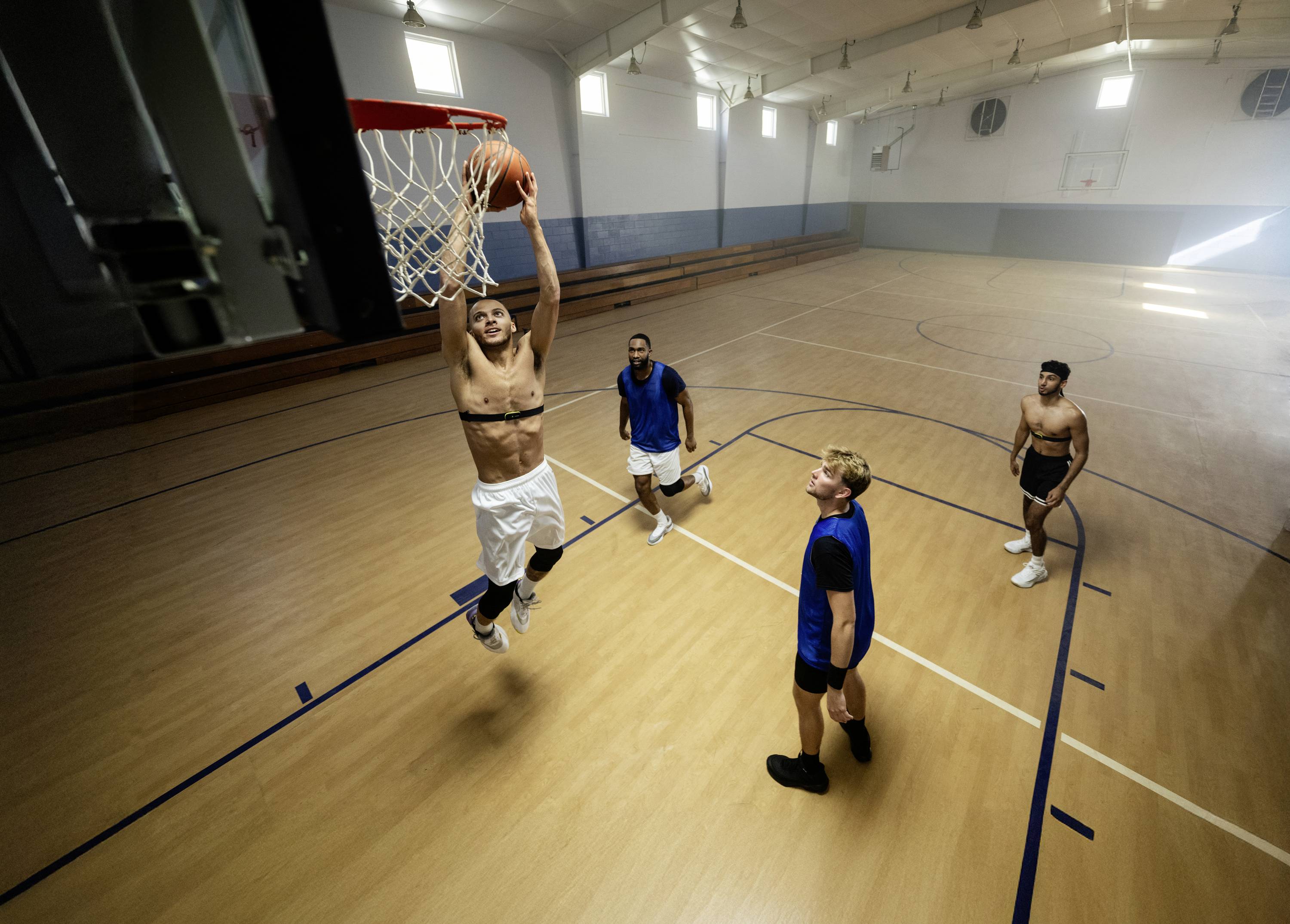 Un giocatore di basket salta per schiacciare, mentre altri tre giocatori guardano. Si trovano in una palestra vuota.