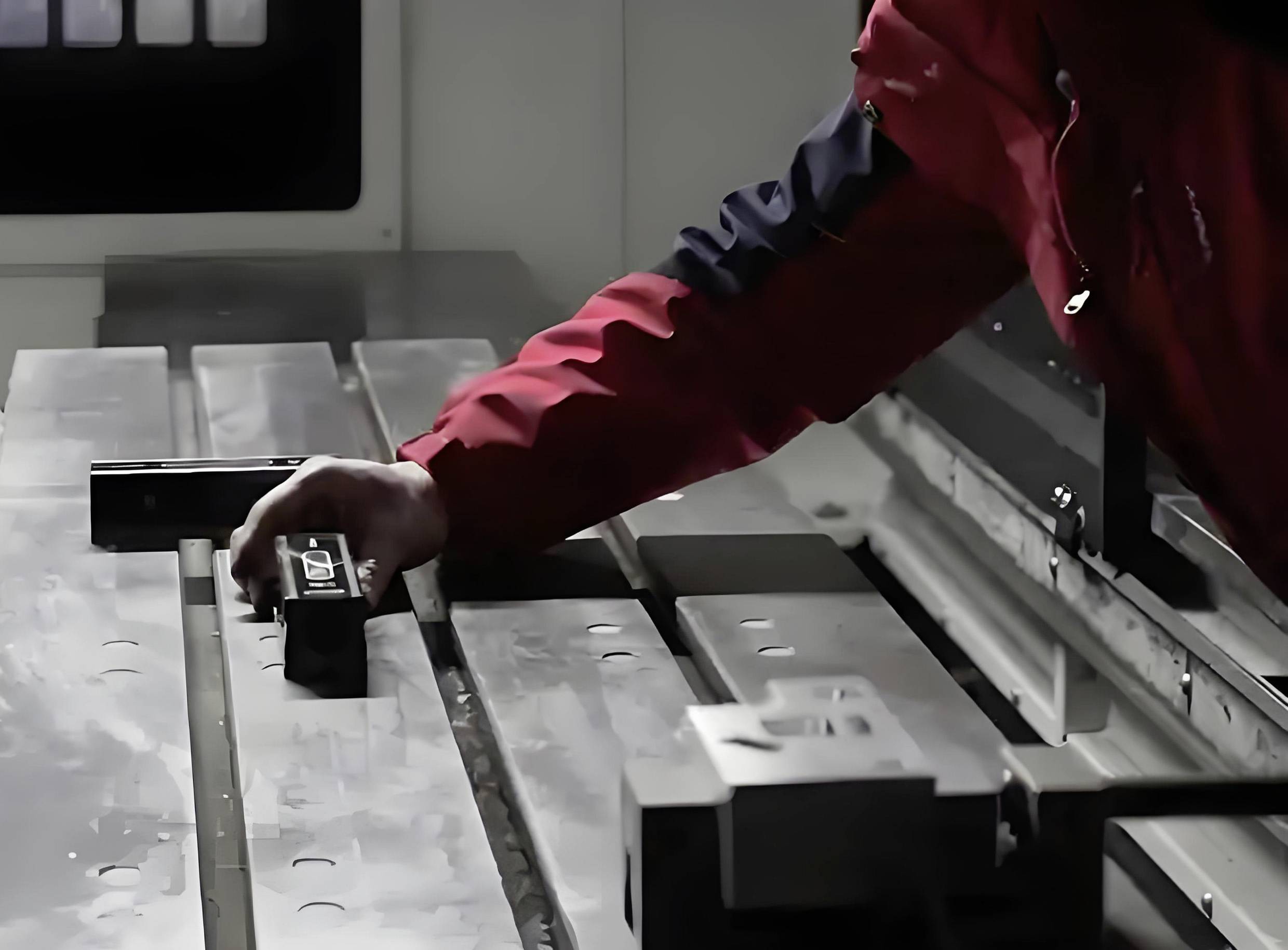 Una persona con una giacca rossa aziona una macchina per la lavorazione dei metalli, regolando un componente su un tavolo metallico all'interno di un ambiente industriale.