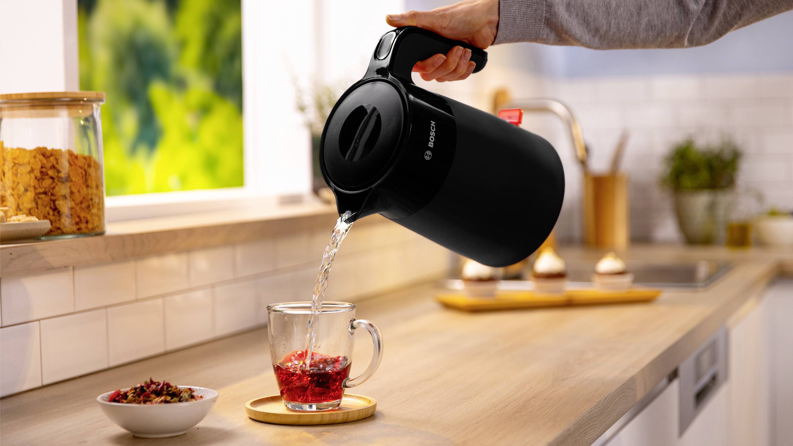Una persona versa acqua calda da un bollitore in una tazza di vetro con erbe su un piano di lavoro della cucina, accanto a una finestra e un barattolo di cereali.